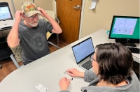 Patient Ron Cleaver puts on hearing aids as audiologist watches on